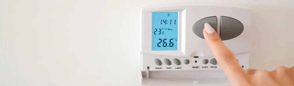 Thermostat blanc sur un mur blanc, affichant 14:11 et 26.6°C. Un doigt appuie sur un bouton gris.