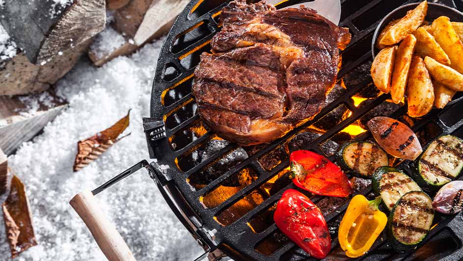 Een barbecuegrill met steak, aardappelen en gegrilde groenten op een besneeuwde ondergrond.