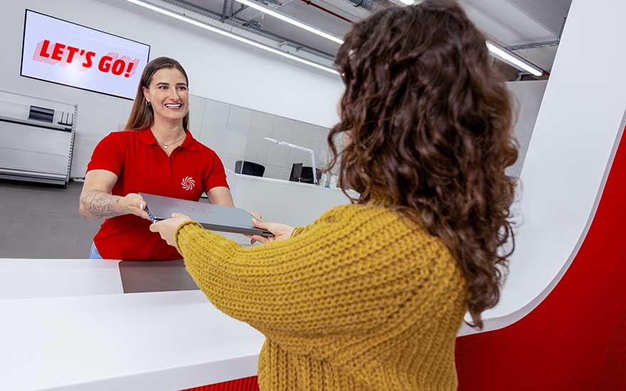 Smiling employee at a white counter hands a laptop to a customer, beside a bright screen displaying the slogan LET’S GO.