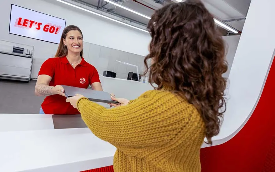 Smiling employee at a white counter hands a laptop to a customer, beside a bright screen displaying the slogan LET’S GO.