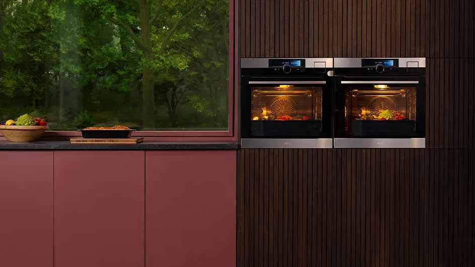 Twee ovens in een houten wand naast een raam met groen uitzicht. Een fruitschaal en bakvorm staan op een rood kastje.