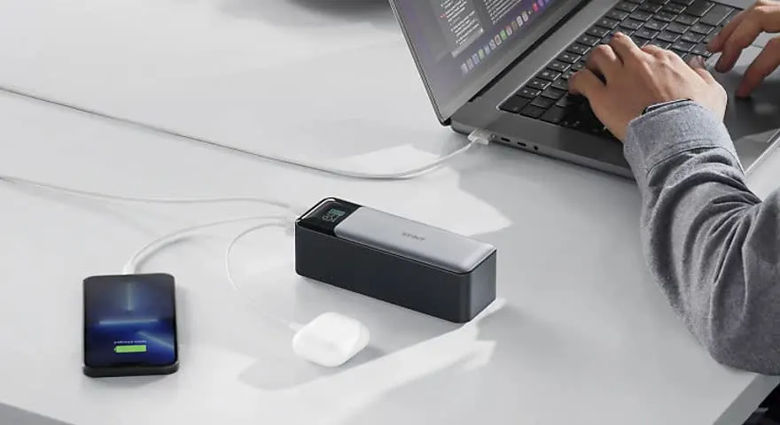 Un ordinateur portable est connecté à un chargeur sur une table blanche, avec un téléphone et des écouteurs à proximité. Une personne tape au clavier.