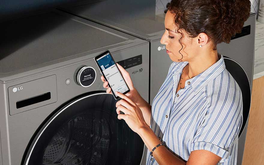 Grijze LG wasmachine en droogkast. Een smartphone toont een app met instellingen.