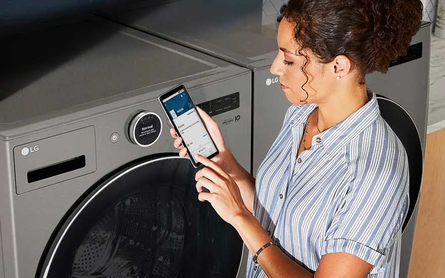 Une machine à laver grise LG est visible. Une main tient un smartphone devant la machine à laver.