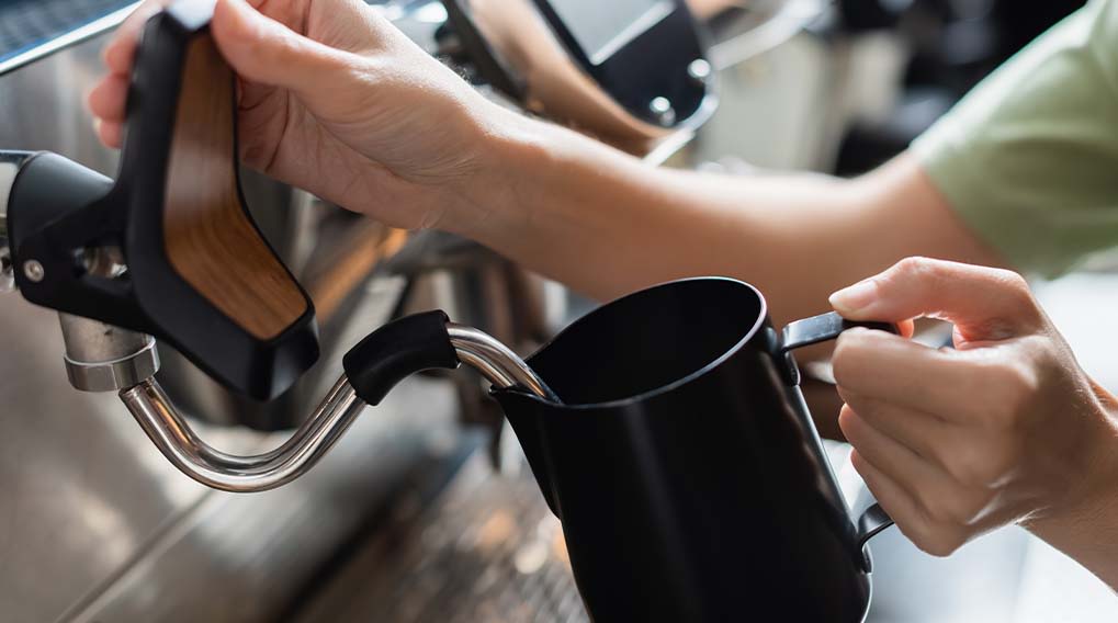 Een persoon giet vloeistof uit een espressomachine in een zwarte kan. De machine is roestvrij staal.