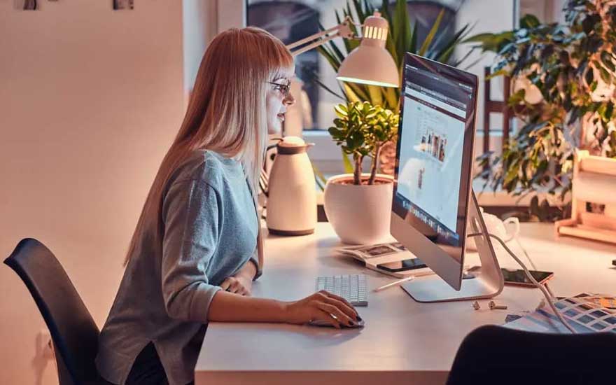 Une femme travaille le soir sur un grand ordinateur de bureau dans un espace chaudement éclairé avec plantes et lampe.