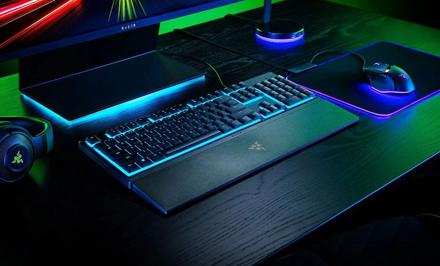 Een gaming setup met een toetsenbord, muis, headset en monitor met blauwe en roze neon verlichting op een donkere houten tafel.