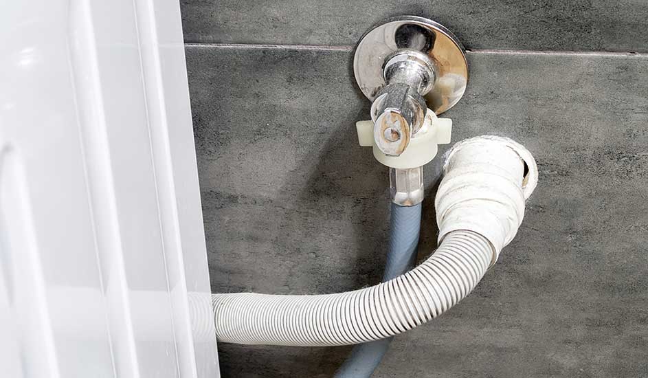 Tuyau blanc raccordé à un robinet chromé sur un mur gris près d'un appareil blanc.