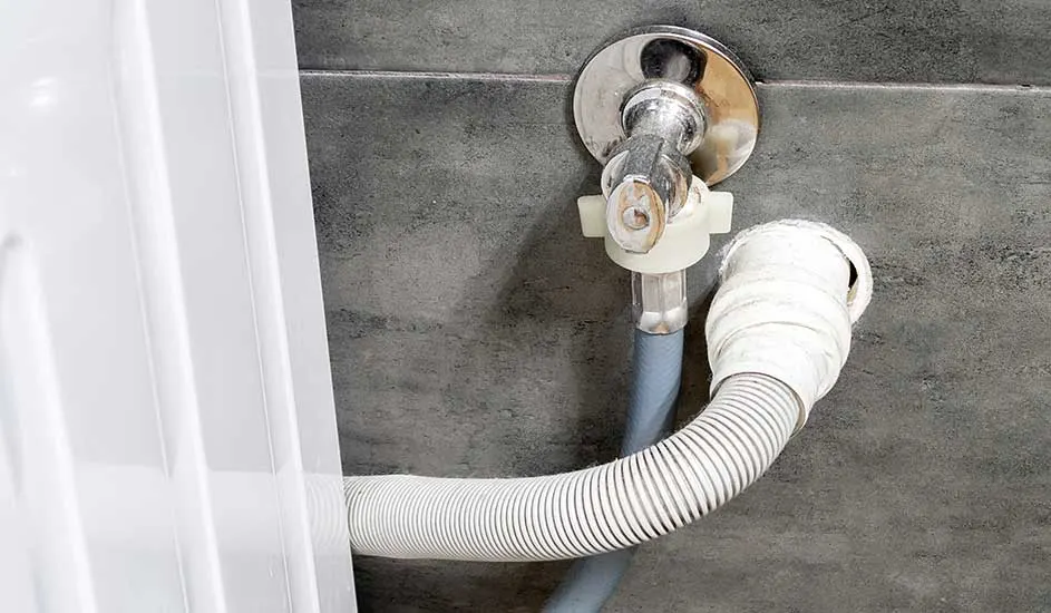 Een witte afvoerslang loopt van een wasmachine naar een kraan op een grijze muur.