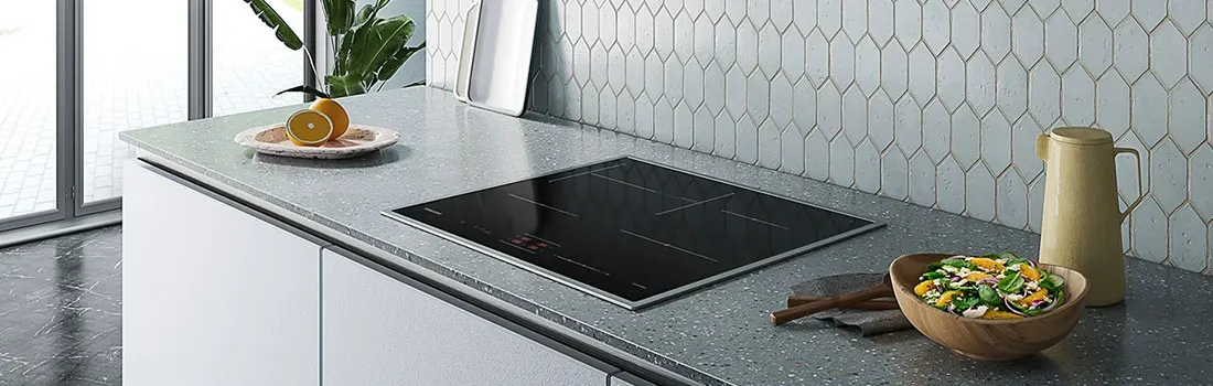Keuken scène: zwarte kookplaat in aanrecht, sinaasappels, salade, en een beige kan tegen een achterwand met zeshoekige tegels.