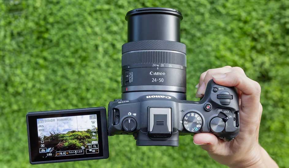 Un appareil photo Canon noir avec un objectif 24-50 est tenu devant un fond vert flou. L'écran LCD affiche un paysage.