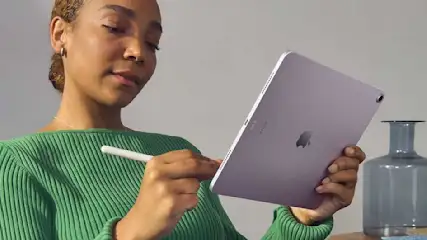 Een paarse iPad wordt met een witte styluspen vastgehouden, groene trui zichtbaar. Grijze vaas op tafel tegen een witte achtergrond.