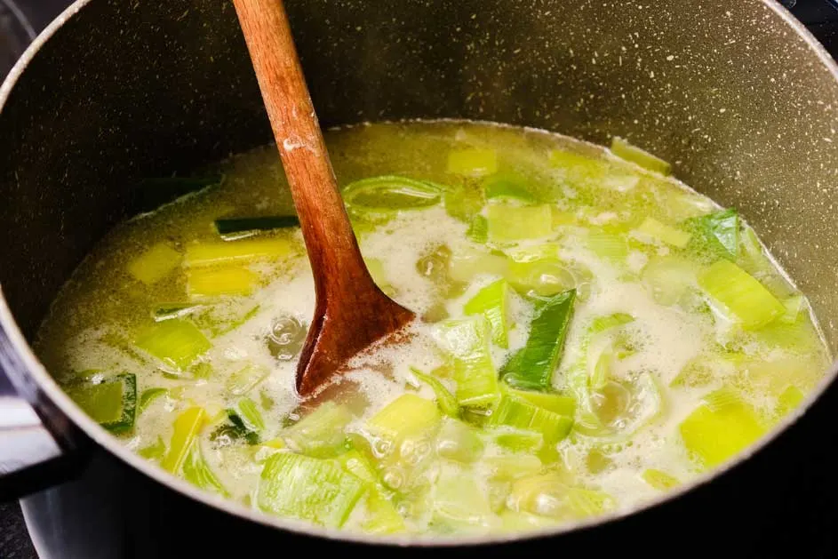 Une casserole contient une soupe avec des légumes verts et jaunes, remuée avec une cuillère en bois.