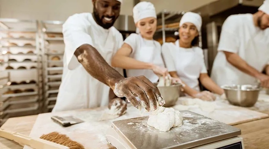 Des boulangers préparent de la pâte sur une table en bois avec une balance et des outils, dans une cuisine professionnelle.
