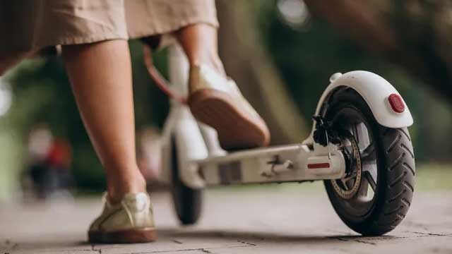 A white electric scooter stands on a pavement, with a foot on it. The background shows blurred greenery.
