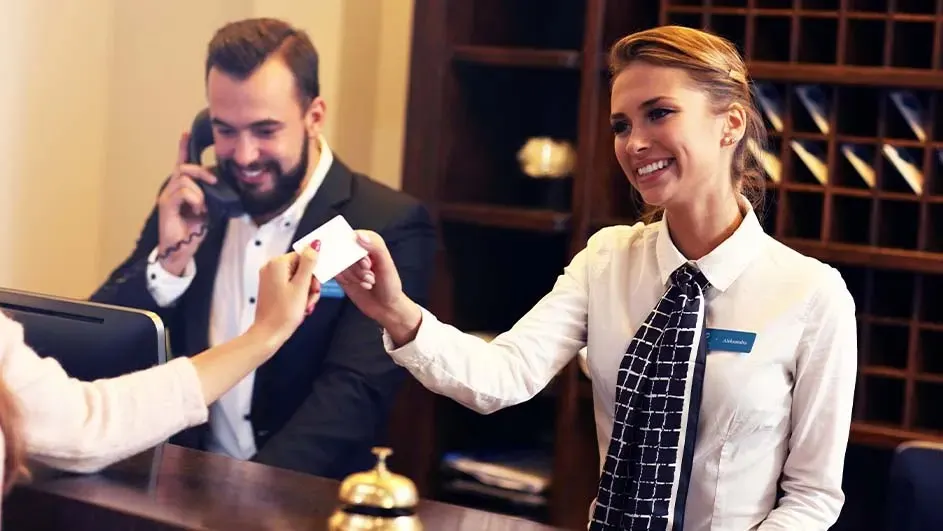 Un comptoir d'hôtel beige avec une cloche dorée. Une réceptionniste en chemise blanche tend une carte. Un homme téléphone en arrière-plan.