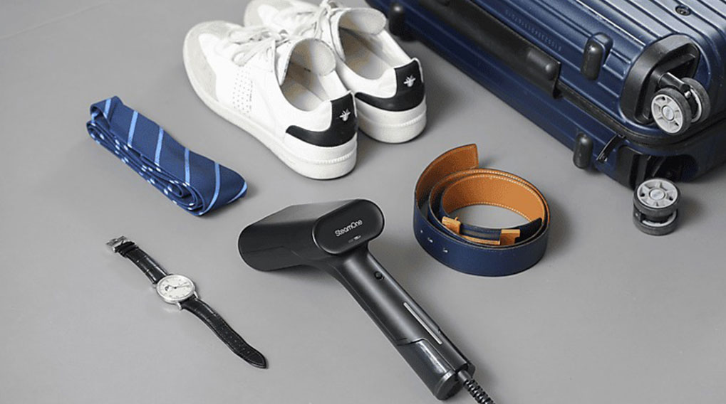 Op een grijze achtergrond liggen witte sneakers, een blauwe koffer, een stoomstrijkijzer, een riem, een horloge en een gestreepte das.