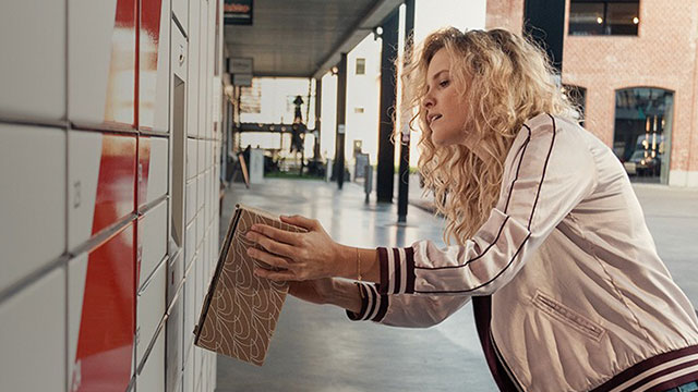 Een beige boek wordt bij een wit en rood locker gehouden. Op de achtergrond is een gang met een gebouw zichtbaar.