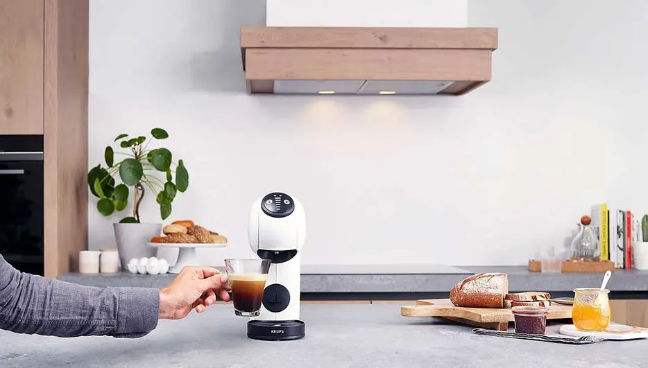 Een witte koffiemachine schenkt koffie in een glas. Op de achtergrond staan brood en planten.