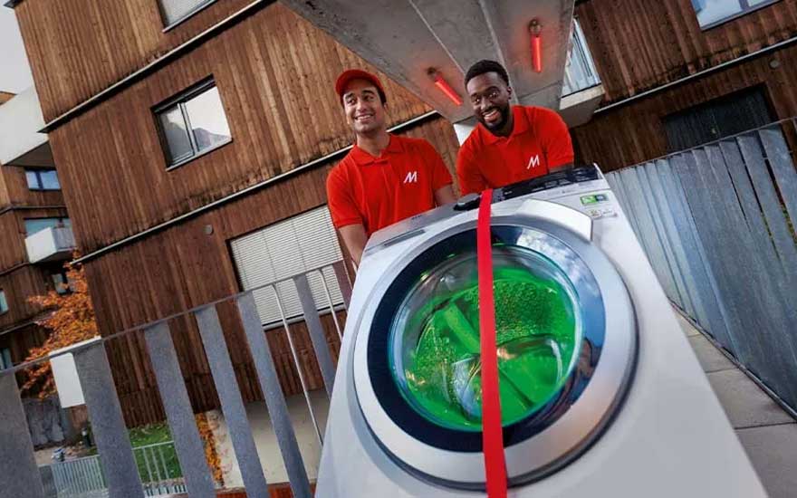 Twee bezorgers duwen een wasmachine met groene vloeistof. Op de achtergrond staat een houten gebouw met ramen en een grijs hek.