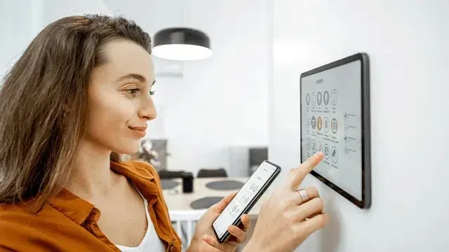 Een vrouw bedient een slim wandpaneel met haar vinger en houdt een smartphone vast. Op de achtergrond staat een helder, modern interieur met lampen.