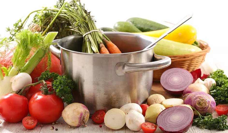Divers légumes et herbes autour d'une casserole en métal avec une louche et un panier. Tomates rouges, oignons violets.