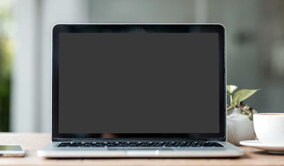 A laptop with a dark screen sits on a wooden table. A smartphone, coffee cup, and plant are nearby. The background is blurred.