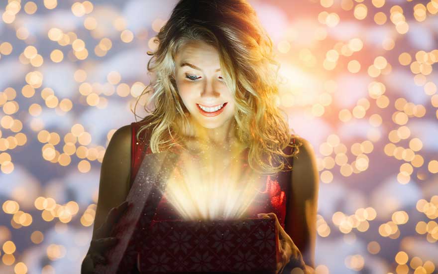 Een verraste jonge vrouw opent een doos waaruit warm magisch licht straalt, omringd door zachte, gouden feestlichtjes op de achtergrond.