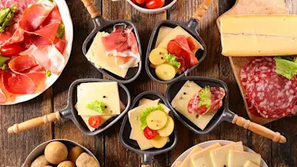 Een tafel met raclettepannetjes gevuld met kaas, aardappelen en vleeswaren. Er zijn ook schalen met ham, salami en kaas.