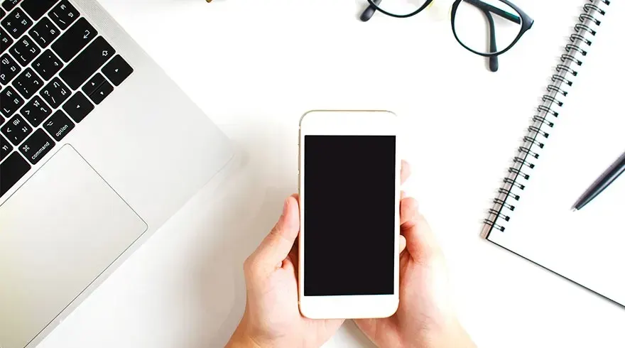 Bureau blanc avec ordinateur, lunettes, carnet, stylo et mains tenant un smartphone noir.