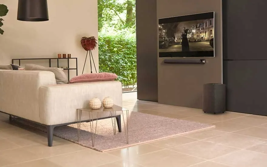 Een woonkamer met een crème bank, een roze tapijt en een tv aan de muur. Buiten is er groen te zien.