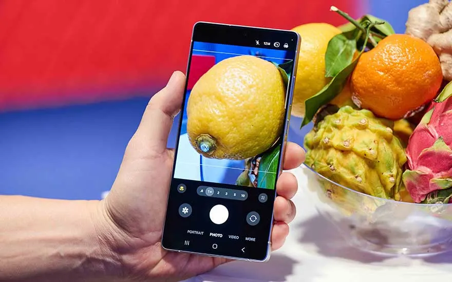 Une main tient un smartphone et photographie un gros citron; à côté, un bol de citrus, gingembre et fruits exotiques, appli photo ouverte.