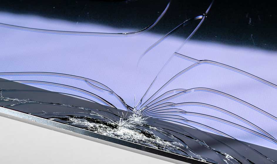 Un écran fortement fissuré repose sur une surface. De nombreuses craquelures partent d’un point d’impact, tandis que des éclats de verre restent visibles.