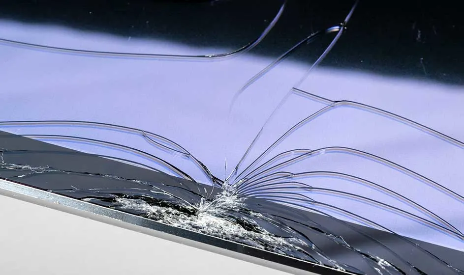 Un écran fortement fissuré repose sur une surface. De nombreuses craquelures partent d’un point d’impact, tandis que des éclats de verre restent visibles.