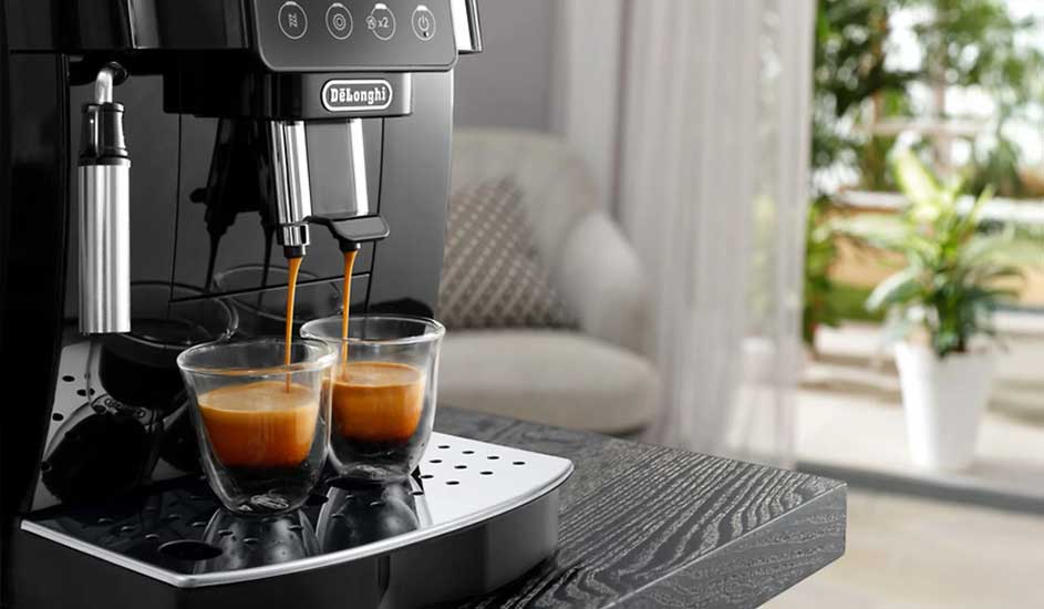 A De’Longhi espresso machine pours coffee into two glass cups. The metal machine sits on a dark worktop, with a plant and chair in the background.