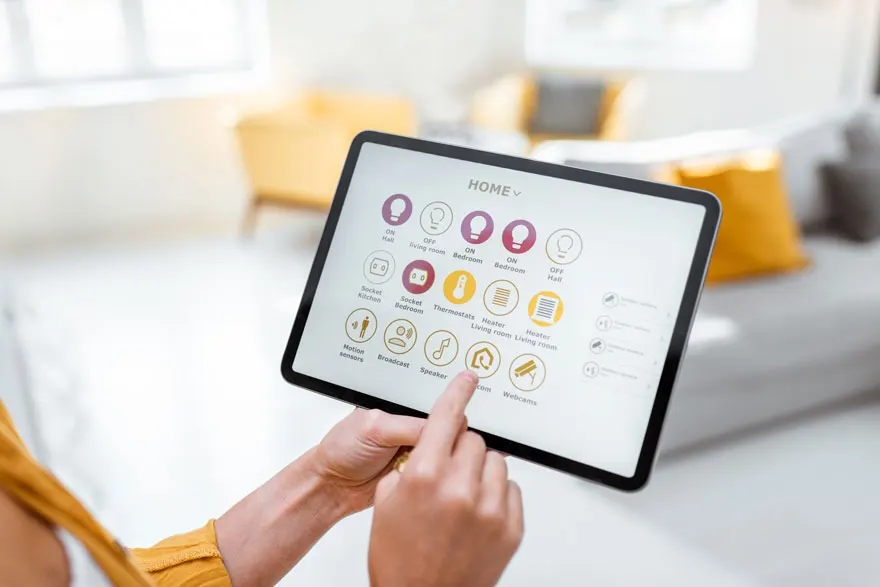 Handen bedienen een tablet met een smarthome-interface in een woonkamer met gele en grijze meubels.