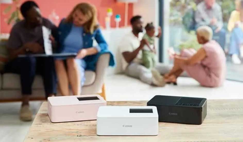 Drie Canon printers (roze, wit en zwart) op tafel. Achtergrond: mensen in een woonkamer.