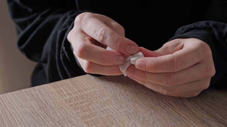 Twee handen manipuleren een wit object boven een houten tafel. Achtergrond: donkere kleding.