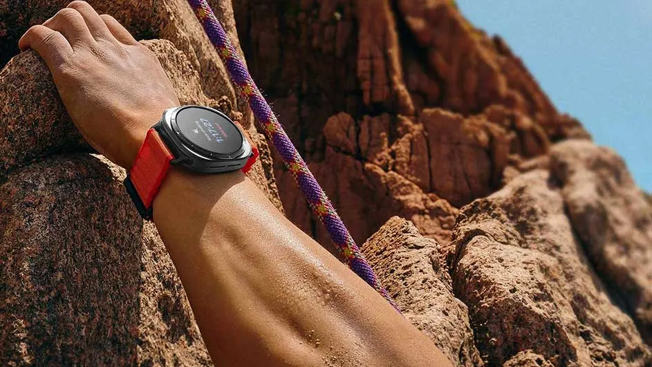 Main tenant un rocher, une montre à bracelet orange est visible. Une corde passe devant la roche brune.