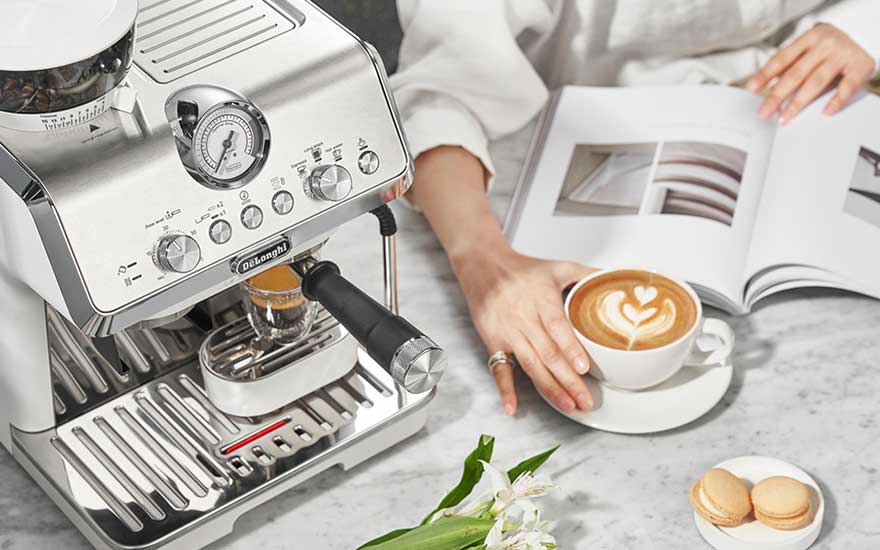 DeLonghi handmatige espressomachine met latte art, naast een open boek en kleurrijke macarons voor een elegant koffiemoment.