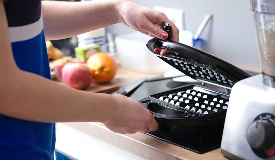 Une personne ouvre un gaufrier noir sur un comptoir de cuisine, avec un mixeur et des fruits en arrière-plan.