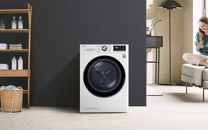 Une machine sèche-linge LG blanche devant un mur sombre, panier à linge et étagère. Une femme est assise sur le canapé avec son téléphone.