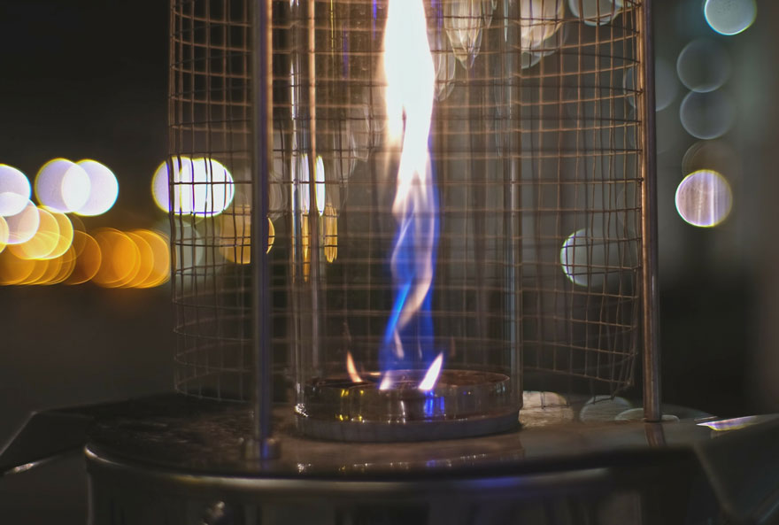 Een gasverwarmer met blauwe vlam achter glas en een metalen rooster. Op de achtergrond zijn vage, gele lichten te zien.