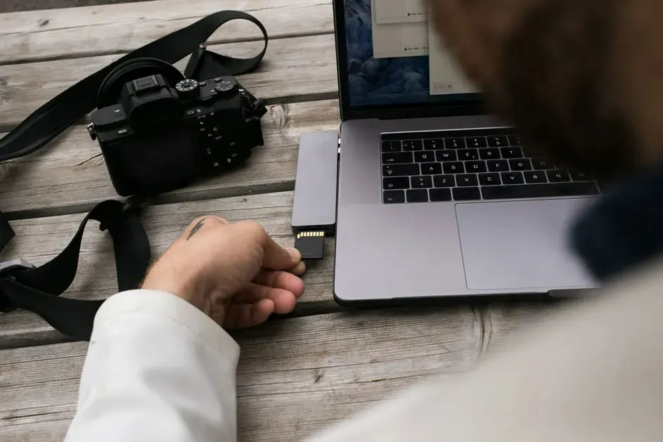 Sur une table en bois, un ordinateur portable gris est connecté à un adaptateur. Une carte SD noire est insérée. Un appareil photo noir est à gauche.