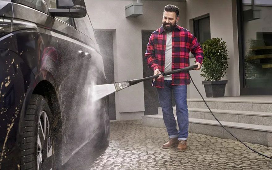 Een bebaarde man in rood geruit overhemd wast zijn donkere auto met een hogedrukreiniger op een klinkerpad voor een modern huis.
