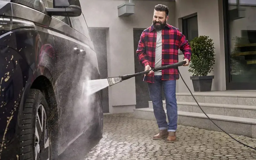 Un homme barbu en chemise à carreaux rouge lave sa voiture sombre avec un nettoyeur haute pression sur un chemin pavé devant une maison moderne.