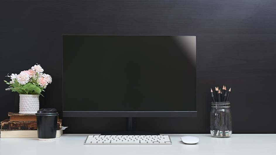 Een bureau met een computer, toetsenbord, muis, bloemen, boeken, een beker en potlood in een pot tegen een donkere achtergrond.