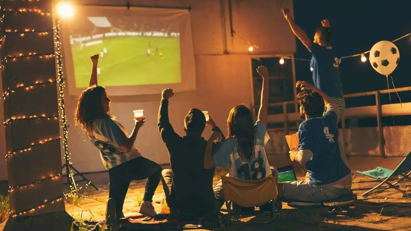 Zes mensen kijken naar voetbal op een groot scherm buiten, sommigen met drankjes in de hand, onder feestverlichting.