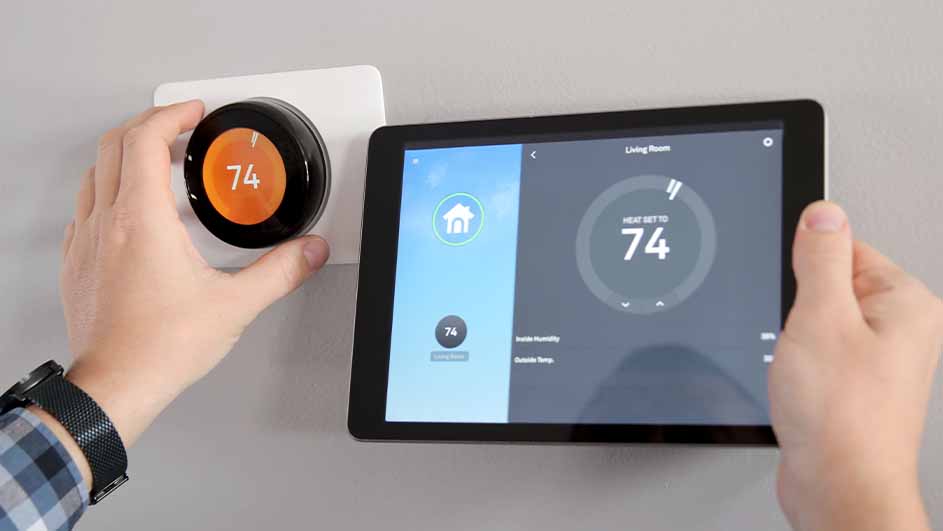 Un thermostat rond noir et orange indique 74, à côté d'une tablette affichant la même température. Le mur est gris clair.