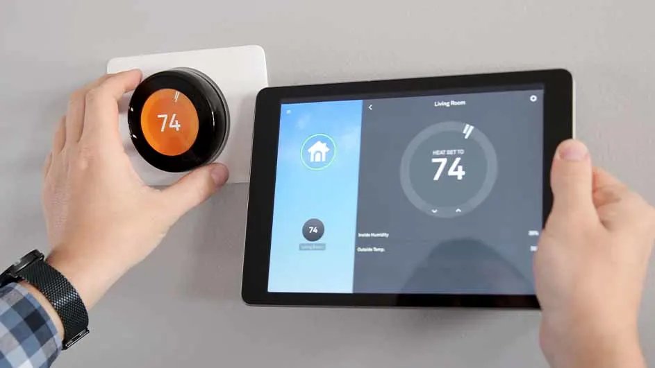 Un thermostat rond noir et orange indique 74, à côté d'une tablette affichant la même température. Le mur est gris clair.